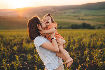 Young mother kissing her baby daughter in the green cornfield on summer day at sunset. Motherhood child care concept. Summer relaxation concept. Summer nature.