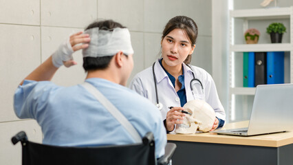 Asian young professional female practitioner doctor in white lab coat with stethoscope holding skull model showing explaining to unrecognizable male patient sitting on wheelchair after brain surgery