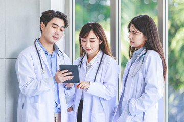 Fototapeta premium Asian young professional male intern practitioner doctor in white lab coat with stethoscope standing holding showing touchscreen tablet computer discussing analyzing with female colleague in hospital