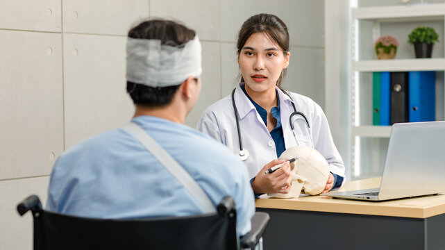 Asian young professional female practitioner doctor in white lab coat with stethoscope holding skull model showing explaining to unrecognizable male patient sitting on wheelchair after brain surgery