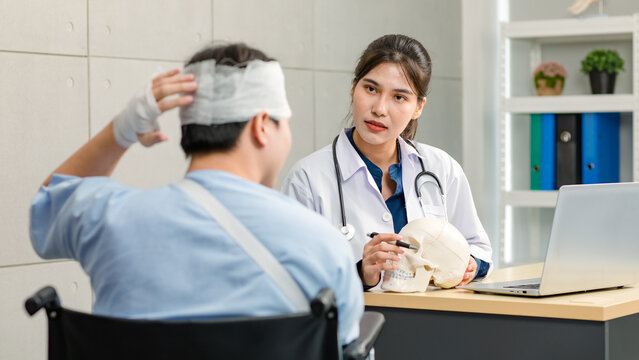 Asian young professional female practitioner doctor in white lab coat with stethoscope holding skull model showing explaining to unrecognizable male patient sitting on wheelchair after brain surgery - Powered by Adobe