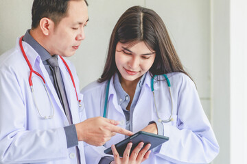 Asian senior professional male mentor practitioner doctor in white lab coat uniform with stethoscope standing helping explaining analyzing data in tablet computer to young female colleague