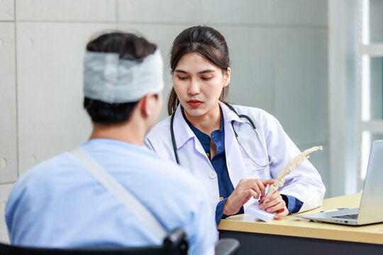 Asian young professional female practitioner doctor in white lab coat with stethoscope holding skull model showing explaining to unrecognizable male patient sitting on wheelchair after brain surgery