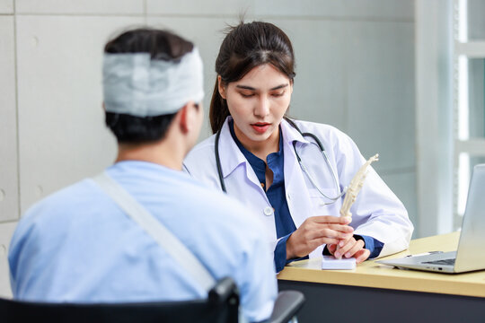Asian young professional female practitioner doctor in white lab coat with stethoscope holding skull model showing explaining to unrecognizable male patient sitting on wheelchair after brain surgery