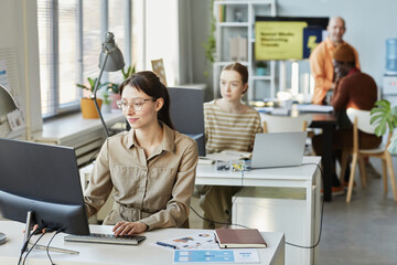 Portrait of young woman using computer at desk in office and smiling with people working in background