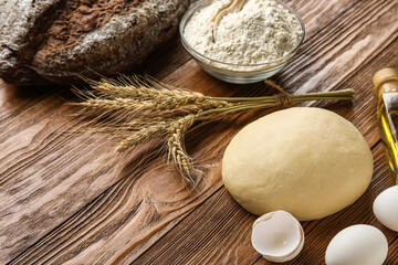 Fresh rye bread, bowl of flour, spikelets, raw dough, oil and eggs on wooden background