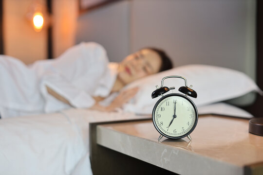 Alarm clock with blurred image of oversleeping woman wearing white sleepwear lying or sleeping on bed in bedroom with happiness and smile. Lifestyle concept