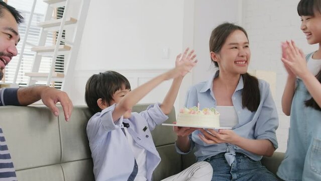 Happy Asian Thai family, young son is surprised with birthday cake, gift, blows out candle, and celebrates party with parents together in living room, wellbeing domestic home event lifestyle.