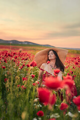 Fototapeta na wymiar Portrait of a woman in a poppy field wearing a wide-brimmed hat at sunset. Vertical photo.