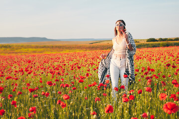 A young woman poses in a field with red wild poppies at sunset. She is holding a glass of strawberries. Front view.