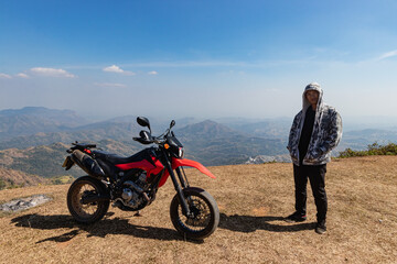 A motorcyclist is standing beside a motorcycle at the top of the mountain. In the distant background are roads and temples. Pha tud, Phetchabun Province, Thailand