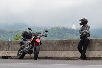 A motorcyclist standing next to a motorcycle The background was a mountain and a white mist.
Park...