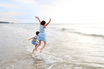 Cheerful Asian child girl and little boy having fun jumping on wave at tropical sand beach at sunset. Happy family enjoy in summer holiday.