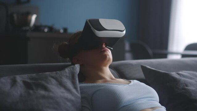 Woman in virtual reality googles enters metaverse immersive expirience via headset interface resting on sofa in minimalistic interior. Girl gaming in cyber space. Futuristic concept.