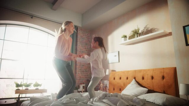 Small Cute Daughter Having Fun at Home with Young Mother. Cheerfully Playing at Home, Jumping on a Bed in Stylish Loft Apartment. Happy Childhood and Motherhood Concept. Handheld Slow Motion Footage.