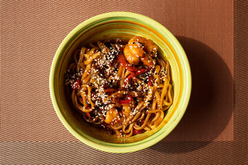 Udon stir fry noodles with vegetables, shrimps and sesame seeds in green bowl on noisy brown table mat background. Top view, close-up