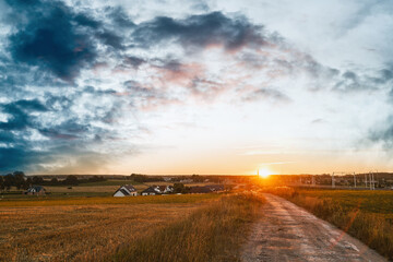 Fototapeta premium Rural country road at the beautiful summer sunset nature landscape. Empty road and tranquil evening scene with warm sunlight. The dramatic sky above the farm.