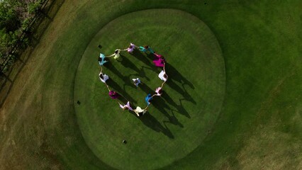 young girls stand in a circle holding hands on green grass in dresses in a group class in nature. Drone view.