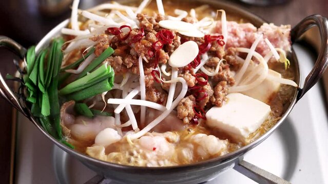鉄鍋で激しく沸騰し、気泡が上がる熱々のモツ鍋。牛モツ、豆腐、ニラ、もやし、挽肉、ニンニクが煮え立ち、湯気が上がる様子を捉えたクローズアップ映像。飲食店メニュー、居酒屋、冬の温かい料理、スタミナ料理の広告素材に。 | Boiling Motsunabe (beef offal hot pot) with vegetables in an iron pot.
