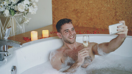 Handsome young man is taking selfie with champagne glass using smartphone in hot tub with foam in...