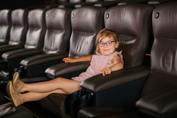 Little preschool girl with glasses watching cartoon movie in cinema and eating popcorn. Happy...