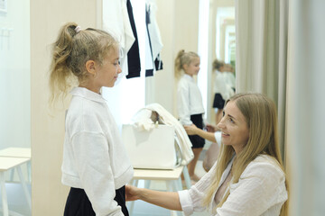 A little girl trying on a jacket and skirt with her mother in the dressing room. Mom buys things for the baby girl. Shopping with children.