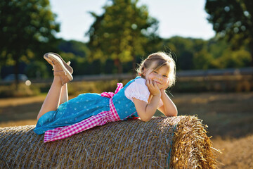 Cute little kid girl in traditional Bavarian costume in wheat field. Happy child with hay bale during Oktoberfest in Munich. Preschool girl play at hay bales during summer harvest time in Germany.