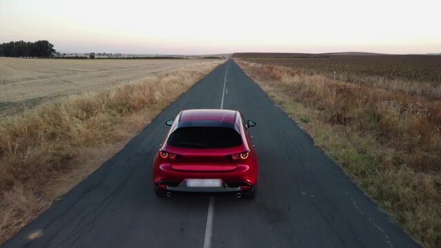 Shiny new red car drives slowly on narrow road through crop fields