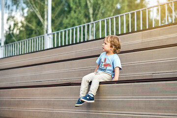 Adorable little boy with funny grimace in stylish casual clothes sit on wooden bench watching movie on huge screen in public park - 526927582