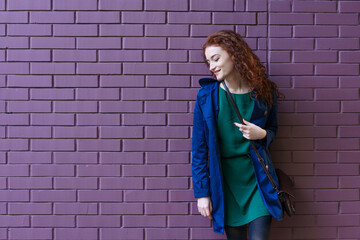 Portrait happy caucasian woman with ginger hair and freckles in stylish clothes, looking at camera posing against lilac brick wall. Concept of youth and happiness