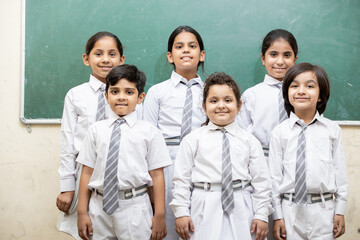 Portrait of happy indian school kids in uniform standing in classroom with chalk board in the background, Elementary school, Education concept.