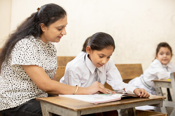 Indian mature teacher helping a student in elementary school lessons in classroom. female child...
