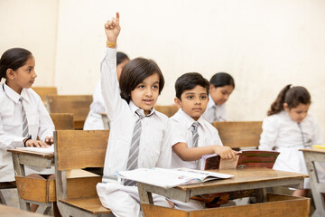 Happy indian schoolboy raising arm to answer a question in the classroom.