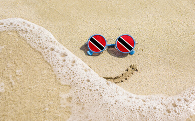 Sunglasses with flag of Trinidad and Tobago on a sandy beach. Nearby is a sea lightning and a painted smile. The concept of a successful vacation in the resorts of Trinidad and Tobago.