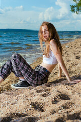Fototapeta na wymiar Blond Caucasian Girl in Casual Clothing Sitting While Resting In Front of Sea Shore At Daytime.