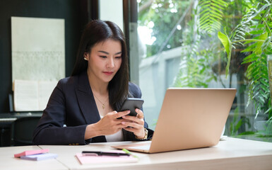 Charming businesswoman sitting in comfortable workplace and using smart phone..