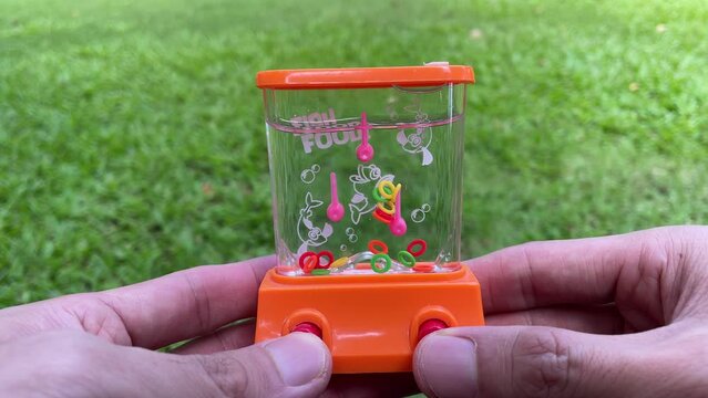 Handheld retro water game ring arcade. Thumb push red button for ring to swirl inside water with nature garden background. Nostalgic fun toy.