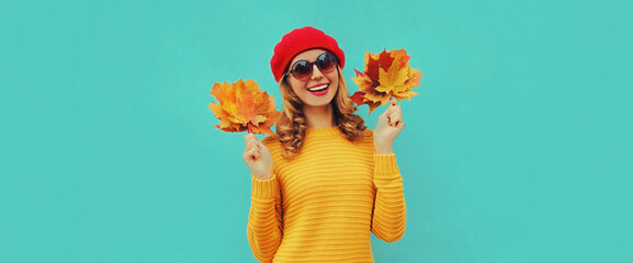 Autumn portrait of happy smiling young woman with yellow maple leaves wearing knitted sweater, red beret on blue background
