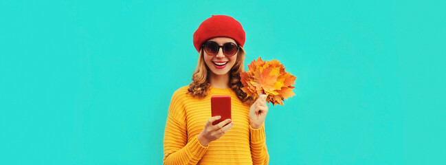 Autumn portrait of stylish happy smiling young woman with smartphone and yellow maple leaves wearing french red beret on blue background - obrazy, fototapety, plakaty