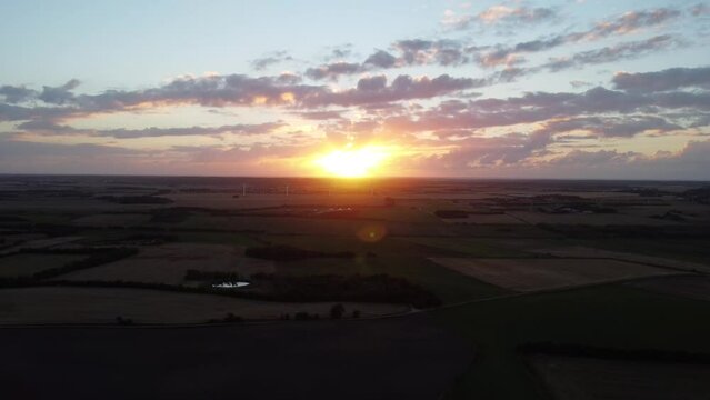 drone flies away from sunset, with lush green farm landscape on the ground, in denmark