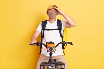 Tired exhausted delivery man in protective mask with thermo backpack on electric scooter, having terrible headache, has flu symptoms, posing with closed eyes isolated on yellow background.