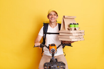 Young smiling courier wearing cap holding pizza boxes and coffee from restaurant or cafe, riding scooter, delivering takeaway food, isolated on yellow background - obrazy, fototapety, plakaty
