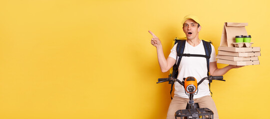 Astonished courier wearing cap and white t shirt holding pizza boxes and coffee, delivering orders on bicycle, pointing at copy space for advertisement, isolated on yellow background