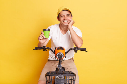 Image of satisfied man wearing white t shirt and cap riding electric scooter and stops to drink takeaway coffee, enjoying his weekend, isolated on yellow background.