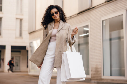 Fashionable young african woman in sunglasses stands on street with bags looking at camera. Brunette wears classic casual clothes. Concept shopping time.