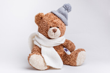 a teddy bear with a thermometer and a knitted scarf and hat is sick on a white background, isolated