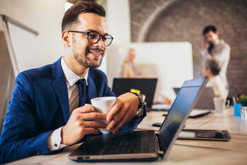 Happy businessman using laptop with team discussing project in the background.