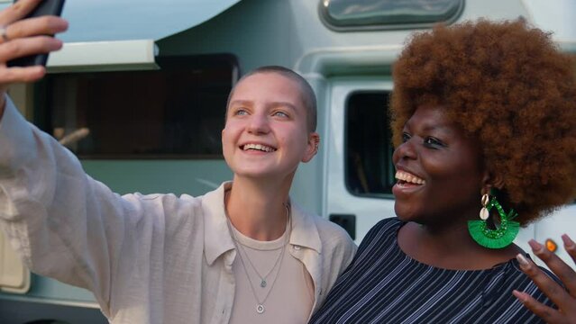 Interracial friendship. Two young diverse girls taking a selfie on a phone together near their camper van. The boys sitting on the stairs and playing the guitar in the background. Reunited on their