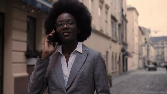 Beautiful african woman in stylish business suit talking on mobile while walking on city street. Successful business lady having conversation on smartphone outdoors.