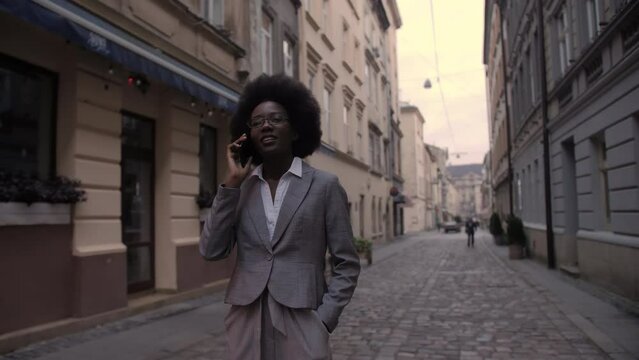 Beautiful african woman in stylish business suit talking on mobile while walking on city street. Successful business lady having conversation on smartphone outdoors.